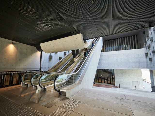 A set of escalators leading to an upper level in the M+ building. They are accessible from an outdoor water promenade. An entrance to the building is visible in the background.